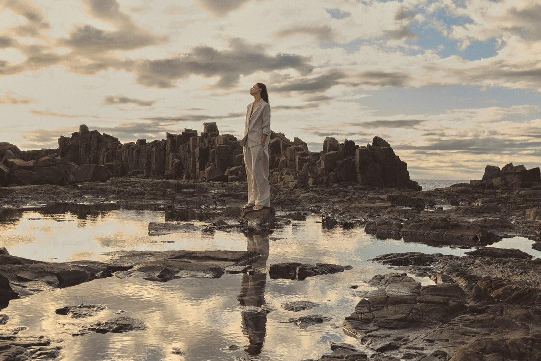 Woman in a cream blazer and trousers standing on coastal rocks beside a reflective pool at dawn