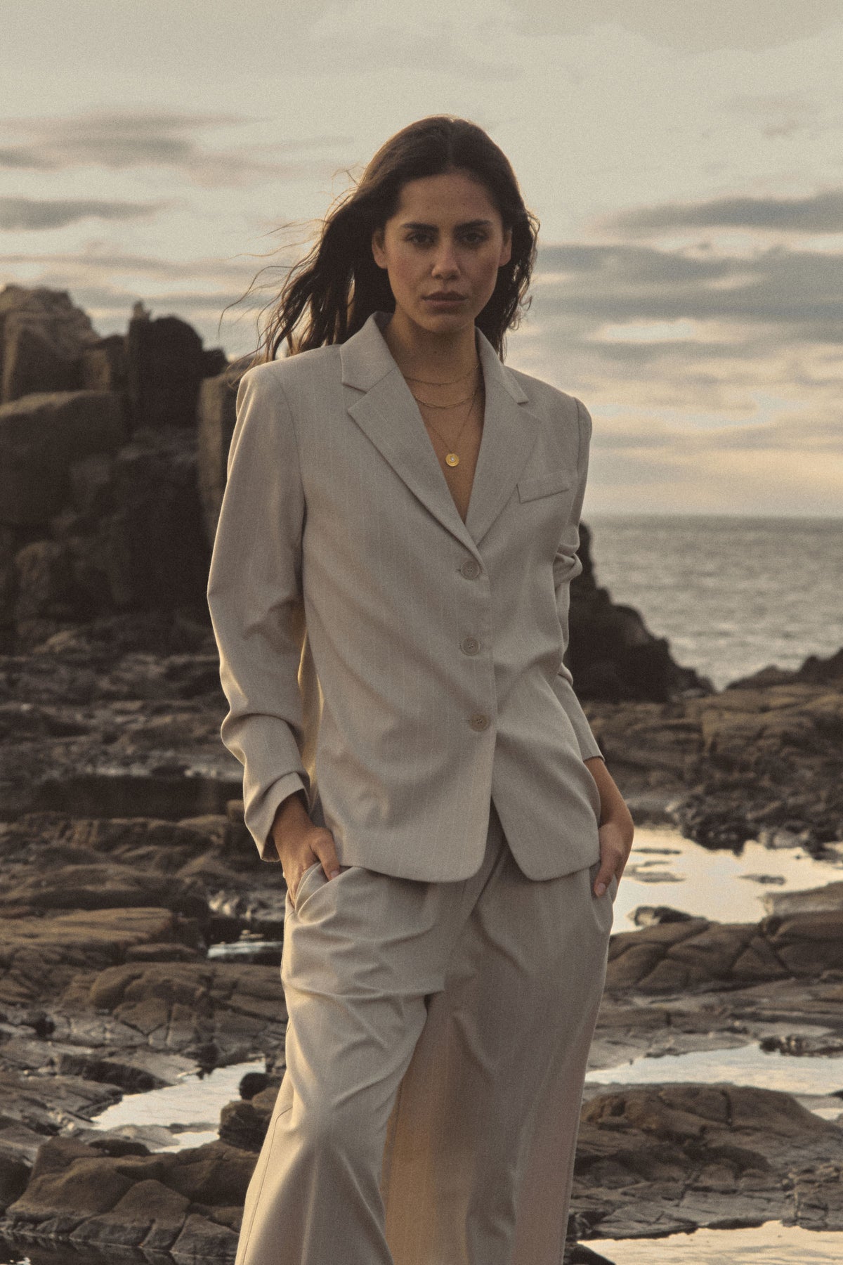 Zoe Relaxed Pinstripe Blazer in beige pinstripe worn with matching trousers on a rocky coastline