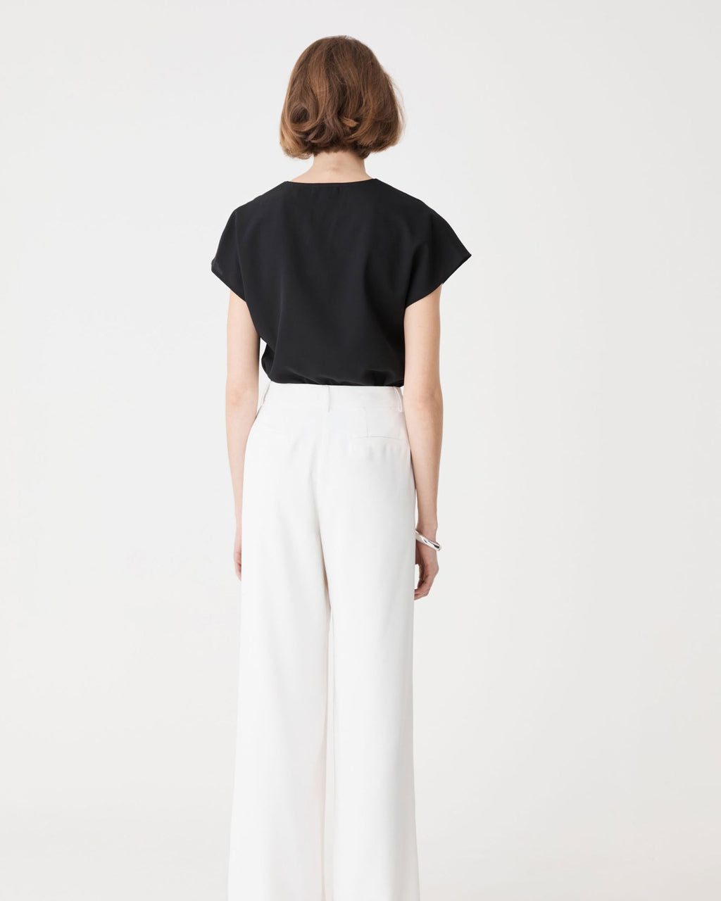 Back view of the black top with extended cap sleeves, worn with white trousers